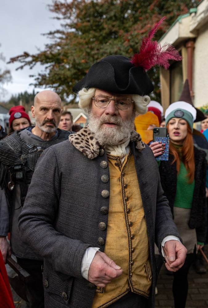 Still from The Fall of Douglas Weatherford, credit Saskia Coulson, which shows Kenneth (Peter Mullan) standing infant of a crowd of people, some of whom are wearing 18th century costumes.
