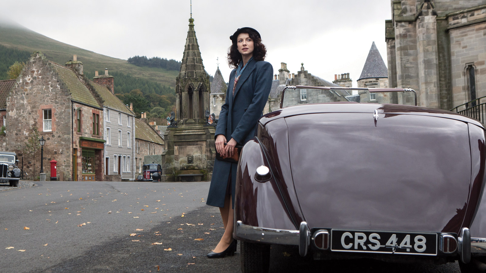 Outlander - Catriona Balfe stands wearing 1940s clothing leaning against a car in a small town